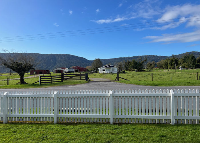 A landscape view of a country road surrounded by green grass and house in the background with a white picket fence in the foreground.