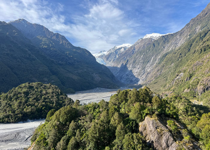 Landscape view of mountains sandwiching a glacier in the background, with large boulders covered by greenery in the foreground.