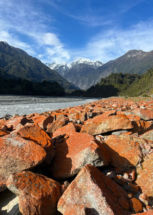 Red algae on rocks appear clearly with green and brown mountain peaks, some snow-capped, in the background.