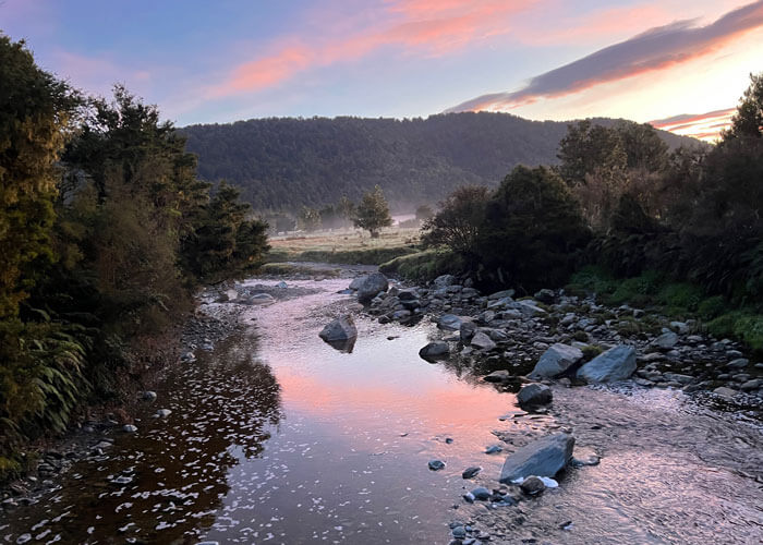 Early-morning landscape view of a waterway winding through a rocky bank flanked by trees, and a forested mounted in the background.