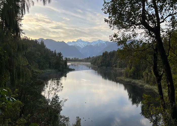 Lake Matheson with trees on either side of the banks features in the middle of the landscape, with the snow-peaked Mount Cook seen in the distance.