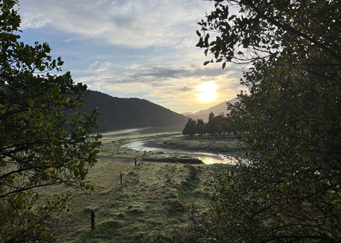 The sun rises in the distance over the silhouette of mountains, with greenery and trees and winding water featured in the foreground.