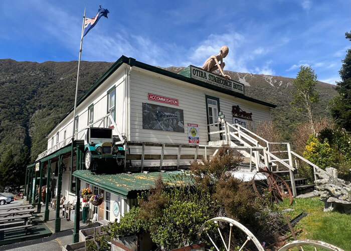 Exterior of Otira Stagecoach Motel, a white, two-story building the stairs leading to the top-story entrance and a large figure of Gollum from the Lord of the Rings stories positioned just above the door looking down.
