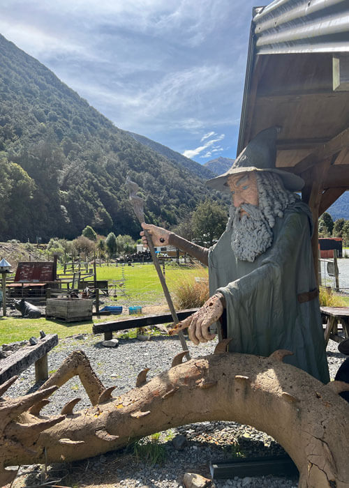 A large statue figure of Gandalf from the Lord of the Rings stories in front of the Otira Stagecoach Motel, with a large tree-covered mountain-range in the distance.