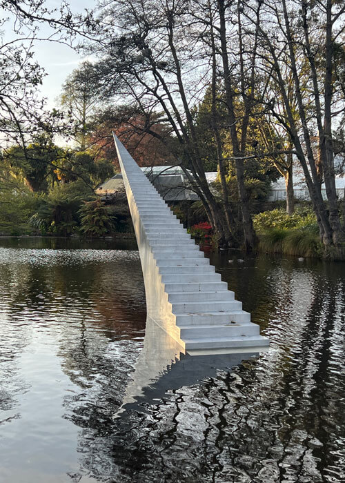 Art of an ascending staircase emerging from the middle of water that is surrounded by trees, shrubs, and greenery.