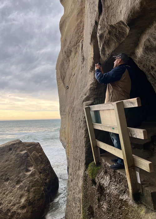 A man wearing a baseball cap and jacket takes a photo from a small outlet looking out onto Tunnel Beach, which is blocked by a wooden railing.