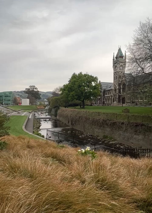 A moat-like waterway winds below and around an old building and clock tower that are surrounded by green grassy landscape and trees.. 