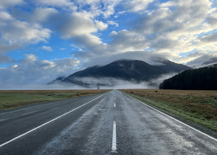 View from the middle of a two-lane highway as fog and clouds loom against blue skies, over and around a mountain in the distance.