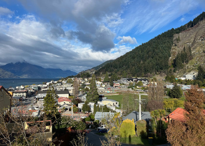 Landscape view of Queenstown, New Zealand with buildings and houses in the foreground and mountains and clouds against a blue sky in the background.