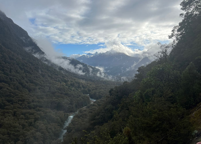Tree-covered Hollyford Valley featured in the center, with mountains in the distance and cloud cover mostly obscuring a blue sky.