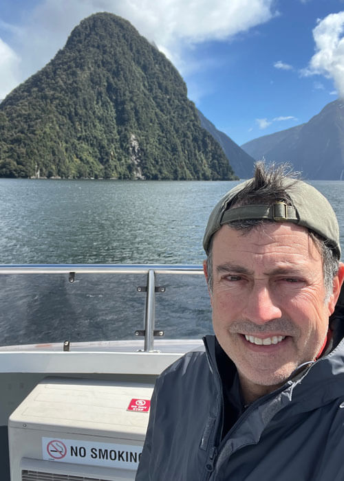 Mark Loftin wearing a gray zipped up jacket and a khaki baseball cap backwards on his head takes a selfie from a boat with the view of tree-covered mountains and water in the background.