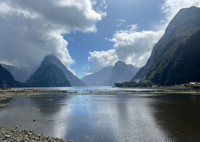 A landscape view of Millford Sound, with tree-covered mountains and water featured against a cloud-filled blue sky.