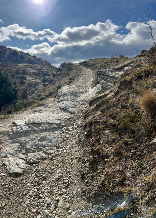 View looking up towards a rocky trail on a mountain, with the sun and clouds showing in the blue sky.